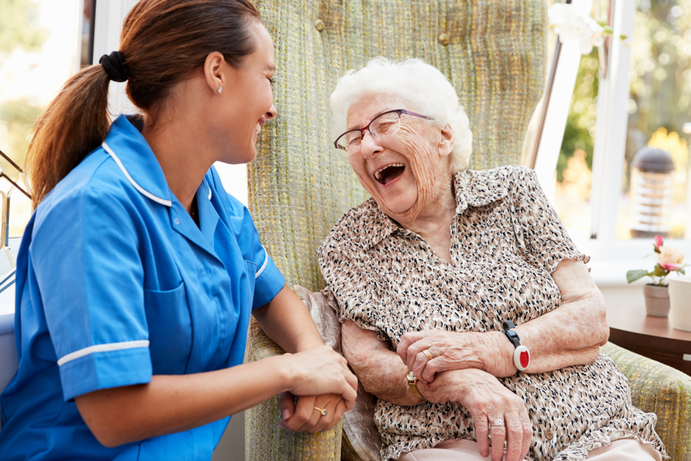 A smiling caregiver in blue talks with a laughing elderly woman seated in a chair.