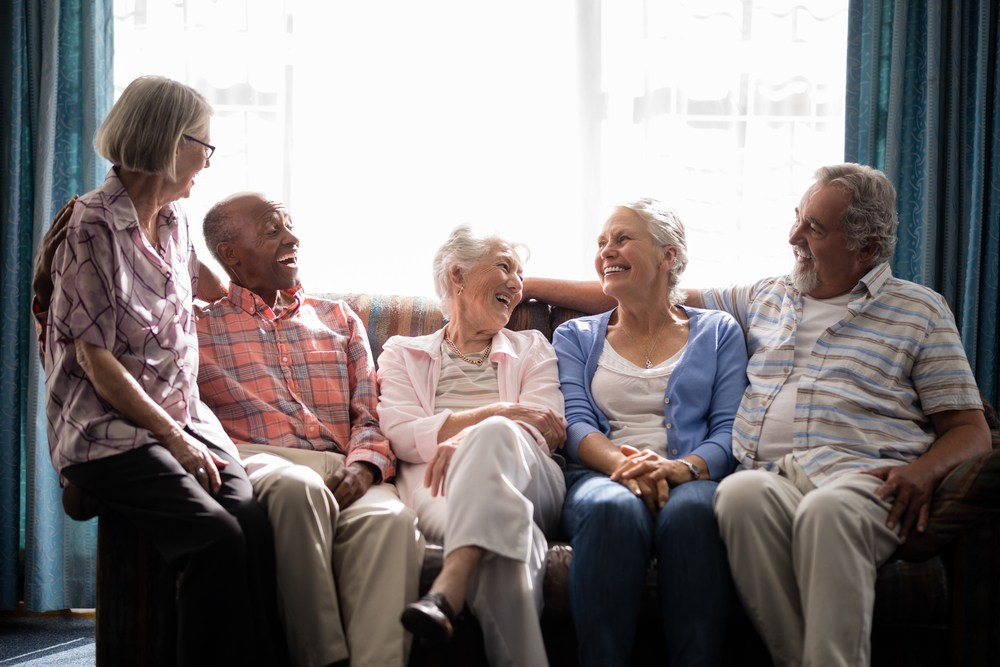 A group of diverse senior friends laugh and talk while sitting together in a bright room.